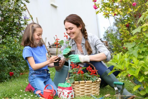 A woman and a young girl are gardening together in a backyard, surrounded by lush green plants, flowering shrubs, and a well-maintained lawn. The girl, wearing a blue T-shirt and pink shoes, is holding a small potted plant, while the woman, dressed in a grey shirt and green gardening gloves, is assisting her and smiling. They are seated on the grass, with gardening tools, a red watering can, and a woven basket nearby, indicating active gardening activities. The garden is vibrant with colorful flowers and dense foliage, set in a natural outdoor environment likely within a residential area in Wood Green. The scene appears to be happening on a bright, sunny day, with clear lighting highlighting the healthy plants and the cheerful interaction. This image exemplifies typical garden maintenance and planting activities that a professional gardening service like Hedge Trimming Wood Green might support, emphasizing lush, well-kept greenery and family-friendly outdoor spaces.