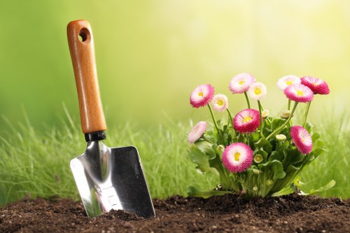 A close-up view of freshly turned dark brown soil in a garden bed, with a small silver hand trowel featuring a wooden handle inserted into the earth. To the right, there is a vibrant cluster of pink and white primrose flowers with rounded petals and green foliage, growing at the edge of the planting area. The background showcases a lush, well-maintained lawn with dense, green grass, softly blurred to emphasize the garden elements in the foreground. The scene appears to be in natural daylight, possibly during a mild weather day, illustrating typical outdoor gardening activity in a residential garden, as seen in areas like Wood Green, suggesting a focus on garden planting and maintenance services provided by companies such as Hedge Trimming Wood Green.