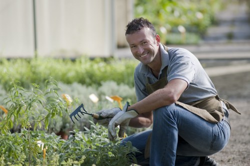 A smiling man dressed in a light grey shirt, dark trousers, and gardening gloves is kneeling in a garden, using a small hand rake to tend to a bed of green plants and flowers. The garden features a well-maintained grassy lawn in the foreground, bordered by a variety of leafy plants and blooming flowers with yellow and white blossoms. Behind him, there is a wooden fence and a backdrop of lush green trees and shrubs, indicating a mature outdoor space in a residential area near Wood Green. The overall setting appears to be a tidy, landscaped backyard designed for outdoor enjoyment and gardening. The lighting suggests a bright, sunny day, creating vibrant natural tones in the scene. This image reflects professional gardening and landscaping activities, such as planting, weeding, and general garden upkeep, which a local gardening service like Hedge Trimming Wood Green could support in maintaining a neat and inviting outdoor environment.