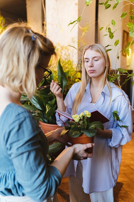 Two women engage in garden maintenance indoors, with one holding a potted plant featuring green leaves and yellow flowers, while the other appears to be inspecting or explaining something about it. Th