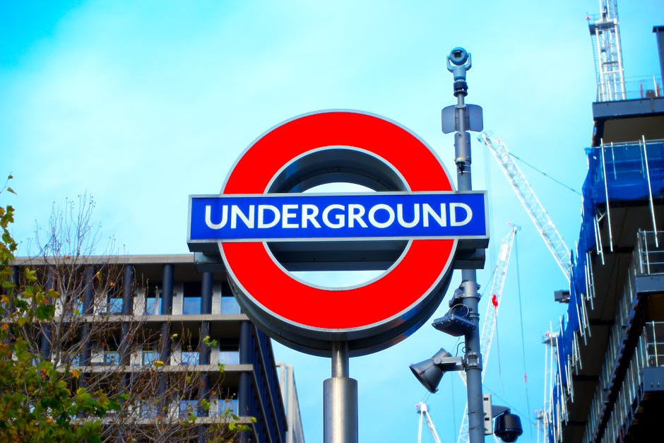 A close-up view of a London Underground sign featuring a circular red ring with a blue bar and white lettering reading 'UNDERGROUND'. Behind the sign, there are modern building facades with balconies 